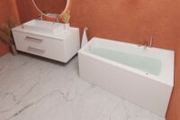 Nemo 160 asymmetrisk badekar i blank akryl. krom armaturer og hvit servant på hvit møbel. Terrakotta vegg og marmorgulv