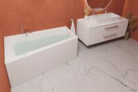 Nemo 160 asymmetrisk badekar i blank akryl. krom armaturer og hvit servant på hvit møbel. Terrakotta vegg og marmorgulv
